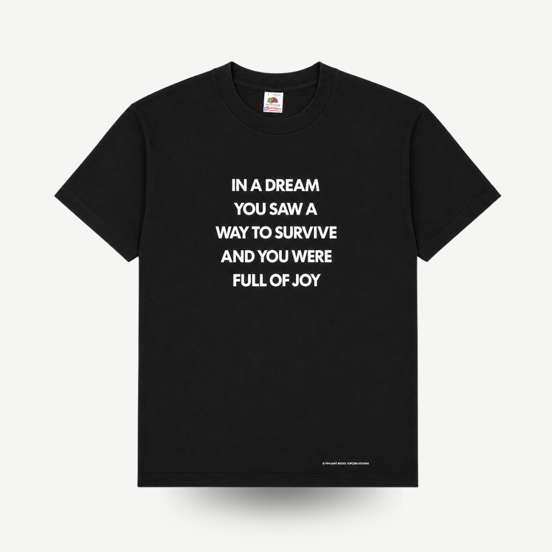 A black short-sleeve crewneck t-shirt with white text on the front reading 'IN A DREAM YOU SAW A WAY TO SURVIVE AND YOU WERE FULL OF JOY'. The shirt is displayed flat against a light gray background with a soft drop shadow.